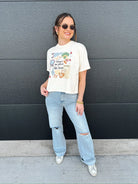 Cream graphic tee with baseball theme "There's no place like home" print, worn by a woman with light wash ripped jeans.