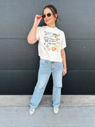 Woman in cream graphic tee with "there's no place like home" baseball design, light wash ripped jeans, and sneakers.