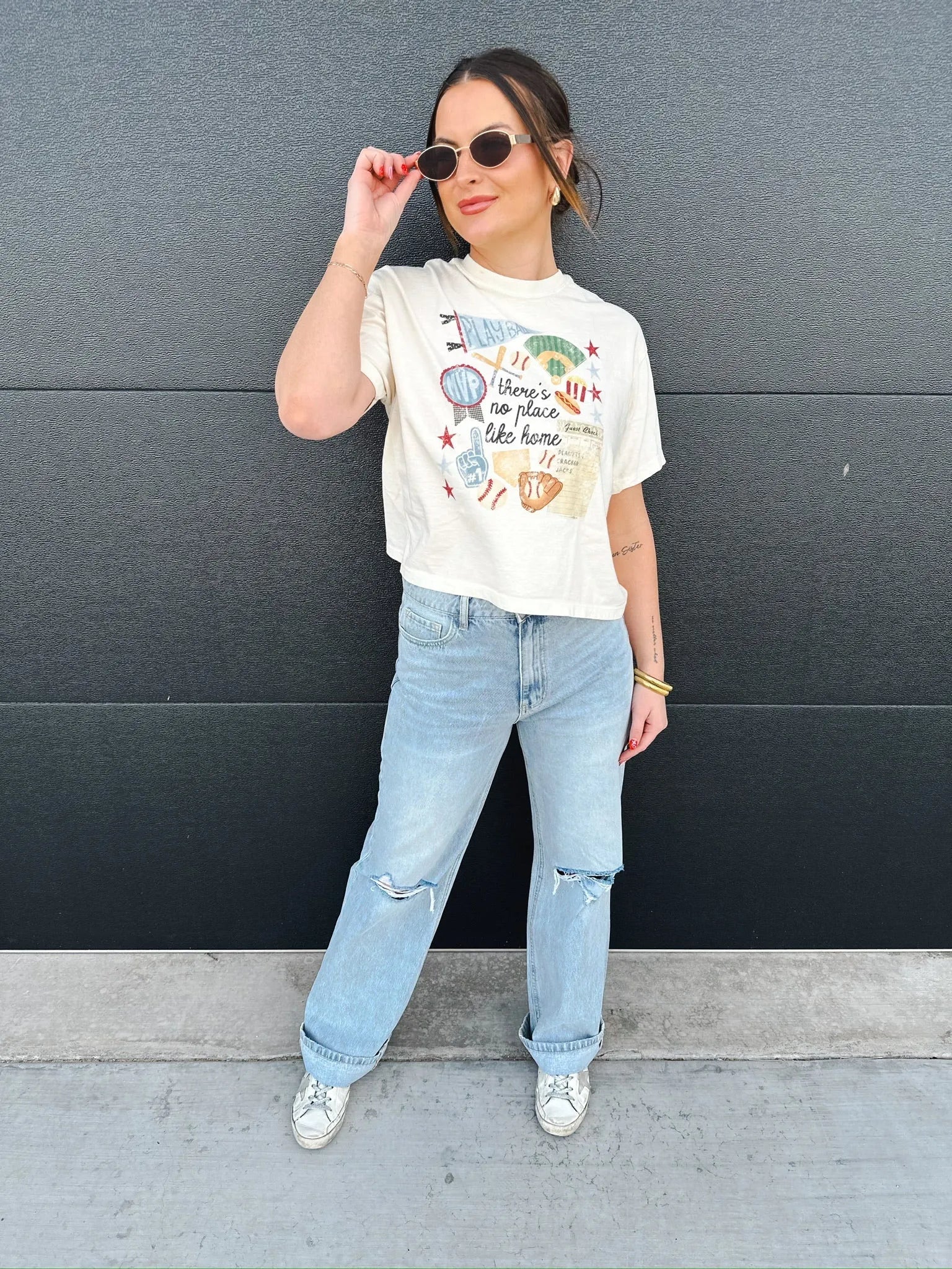 Woman in cream graphic tee with "there's no place like home" baseball design, light wash ripped jeans, and sneakers.
