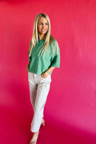 Woman wearing a green button-up sweater and light ecru twill barrel pants with a textured pattern against a bright pink