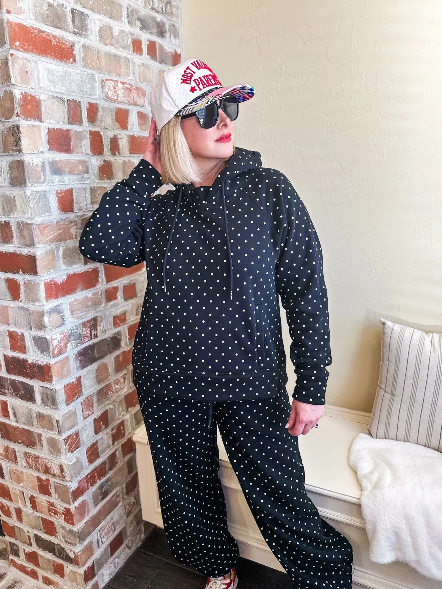 Black and white polka dot sweatsuit with drawstring hoodie and pants. Model wears a baseball cap and sunglasses.