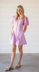 Woman wearing a purple puff sleeve structured mini dress with button front and pleats. She is standing with hands in pockets.