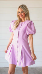 A smiling woman models a light purple mini dress with puff sleeves and a button-down front. The dress features a structured