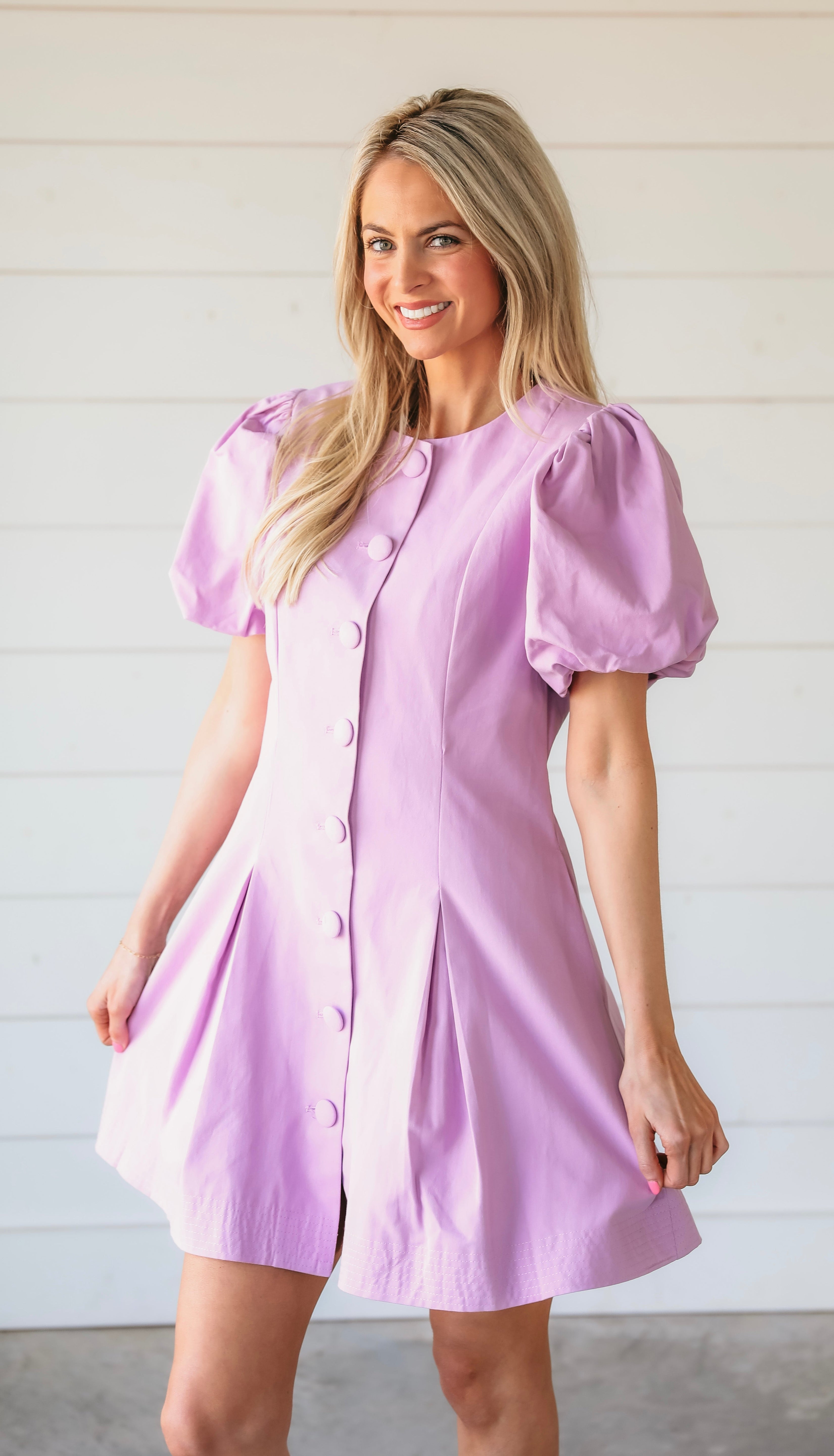 A smiling woman models a light purple mini dress with puff sleeves and a button-down front. The dress features a structured