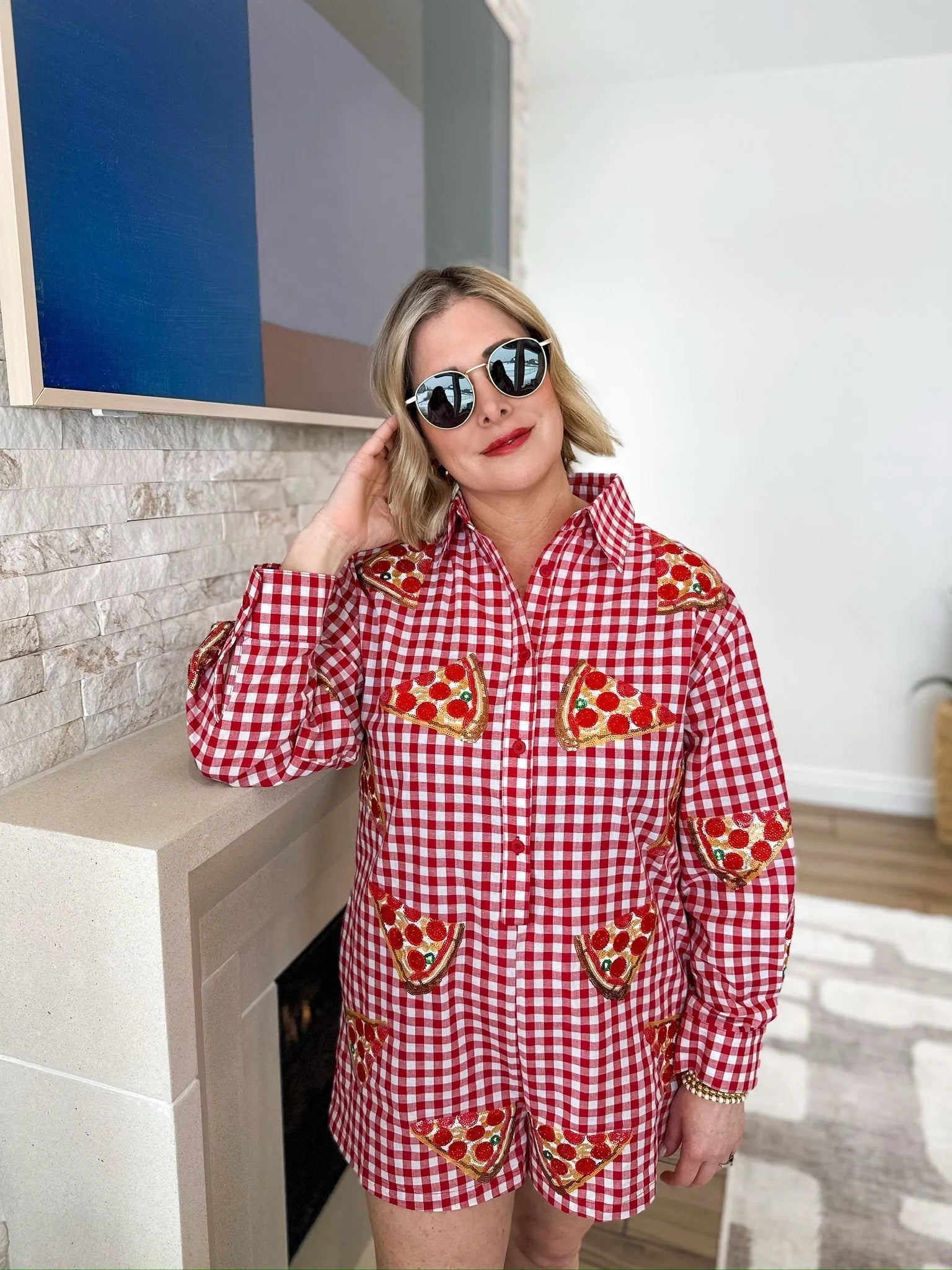 Woman wearing a red and white gingham romper with sequined pizza slices. Round sunglasses, blonde bob haircut.