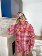 Woman wearing a red and white gingham romper with sequined pizza slices. Round sunglasses, blonde bob haircut.