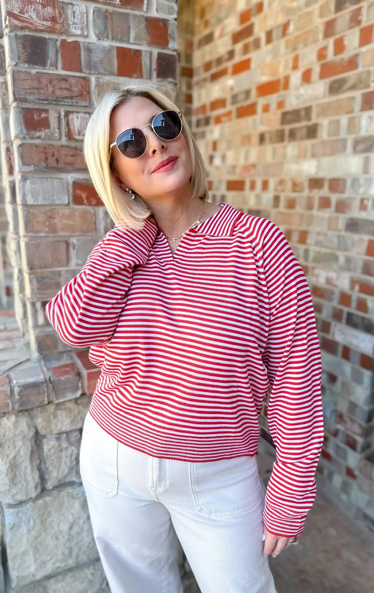Woman wearing a red and cream striped collared sweater pullover top and white pants, accessorized with sunglasses and