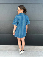 Woman wearing a short-sleeve blue and white striped denim dress with a split neck. She is standing with her back to the