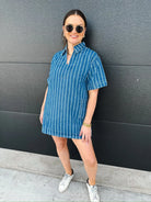 Woman in a relaxed fit short sleeve blue denim split neck dress with white vertical stripes, posing against a dark wall.