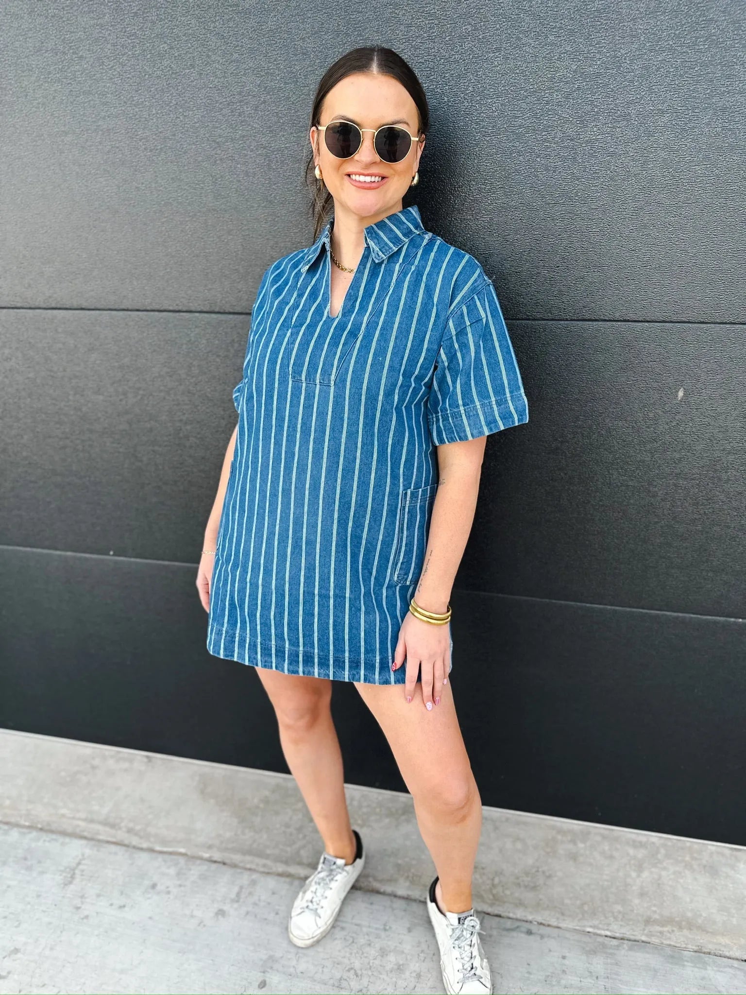 Woman in a relaxed fit short sleeve blue denim split neck dress with white vertical stripes, posing against a dark wall.