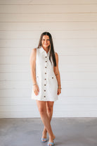 Sleeveless white button-down shirt dress with two chest pockets, a collared neckline, and brown buttons. A woman models the