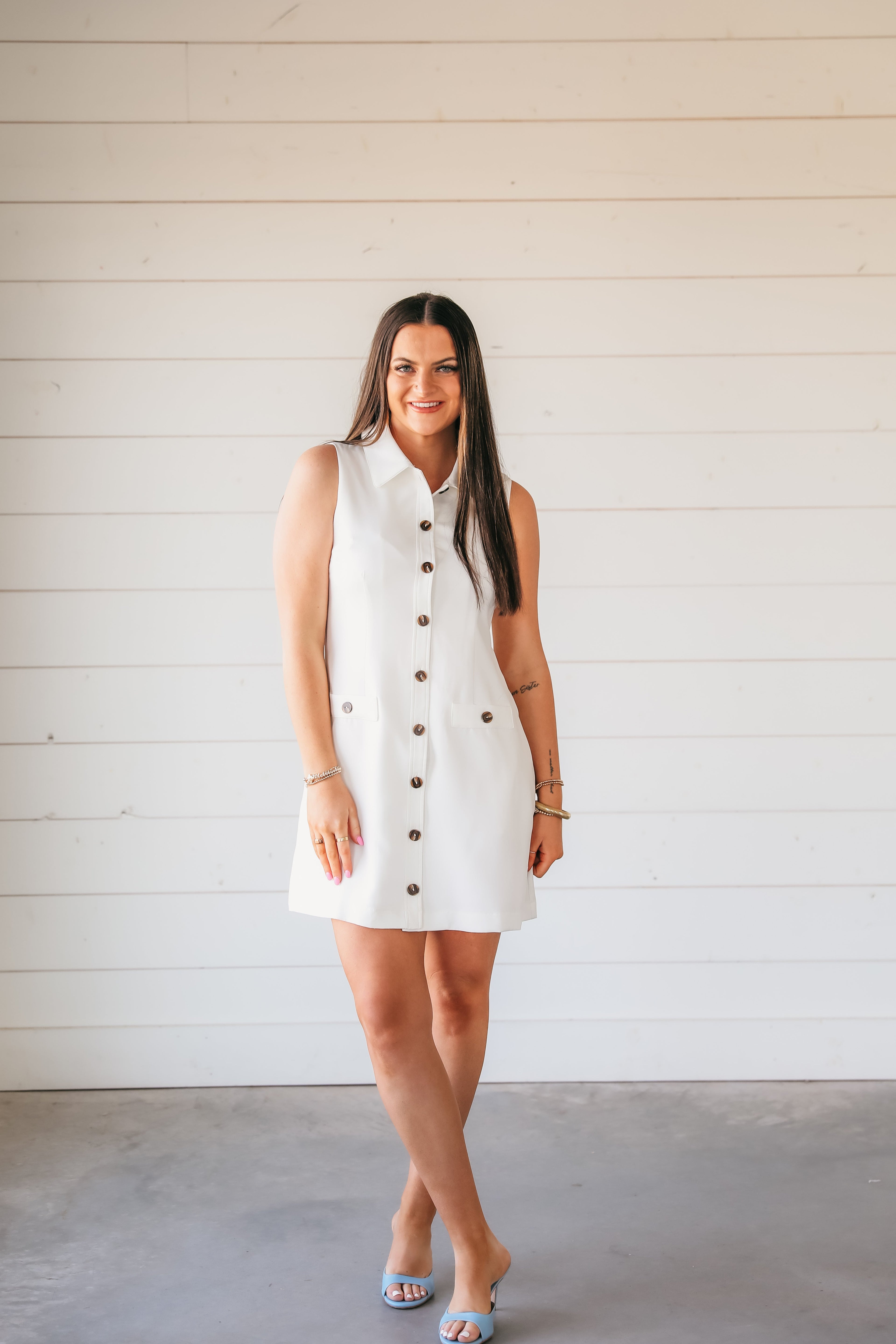 Sleeveless white button-down shirt dress with two chest pockets, a collared neckline, and brown buttons. A woman models the