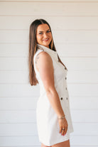 Sleeveless white button-down shirt dress with dark buttons. Model wears bracelet and rings. Clean, modern style.