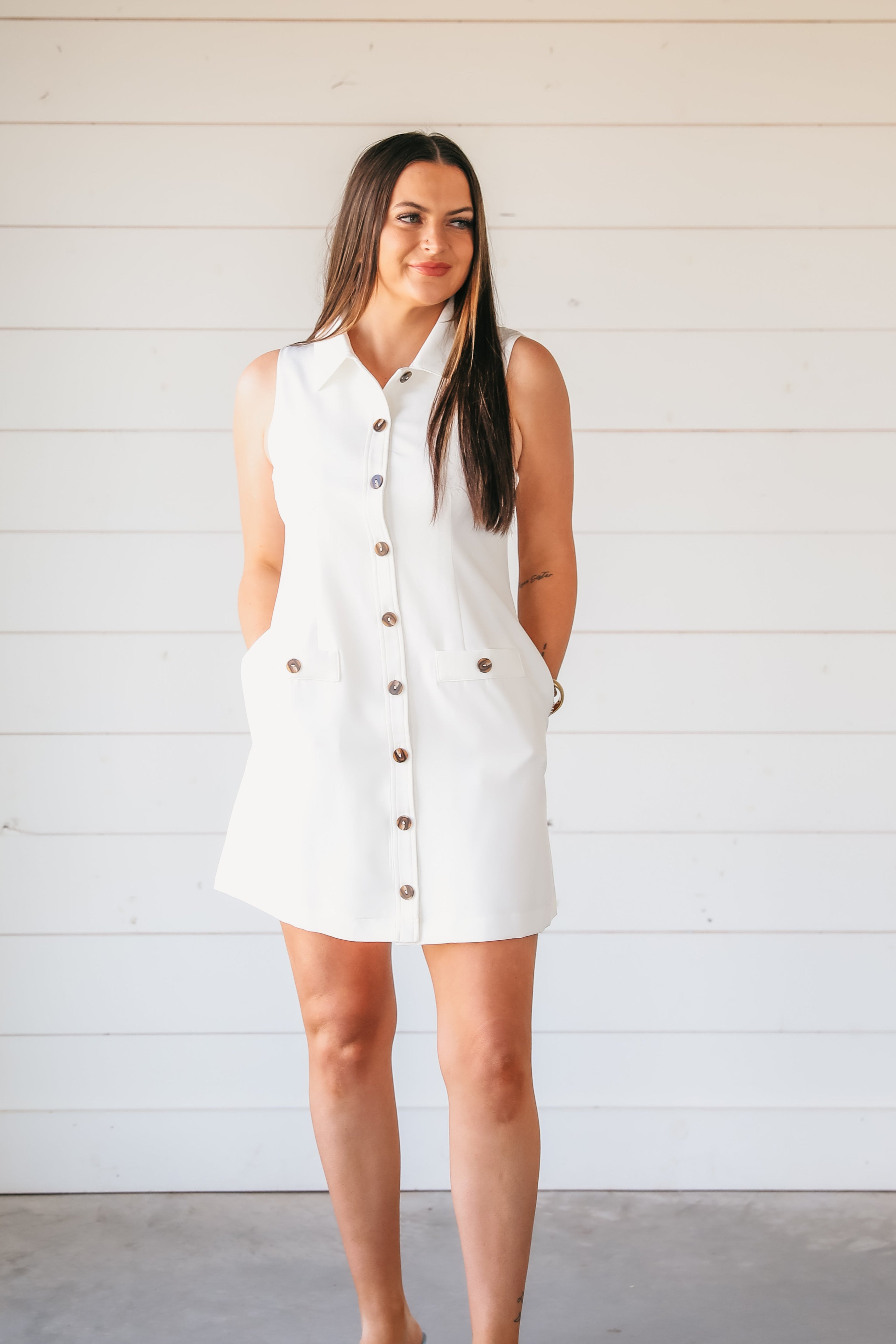 Woman wearing a sleeveless white button-down shirt dress with wood-tone buttons and two chest pockets, standing against a