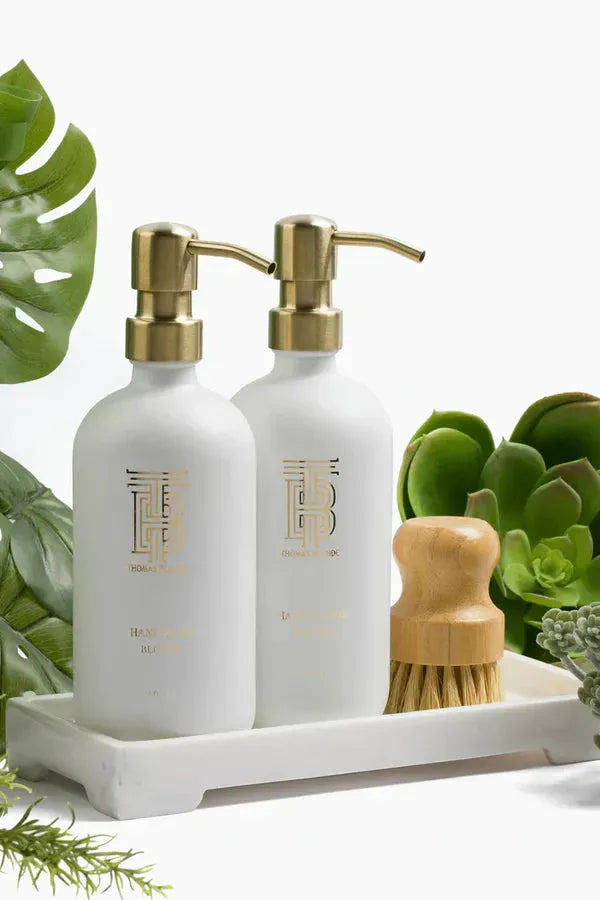White soap and lotion bottles with gold pumps and logos on a white tray next to a small wooden scrub brush, surrounded by