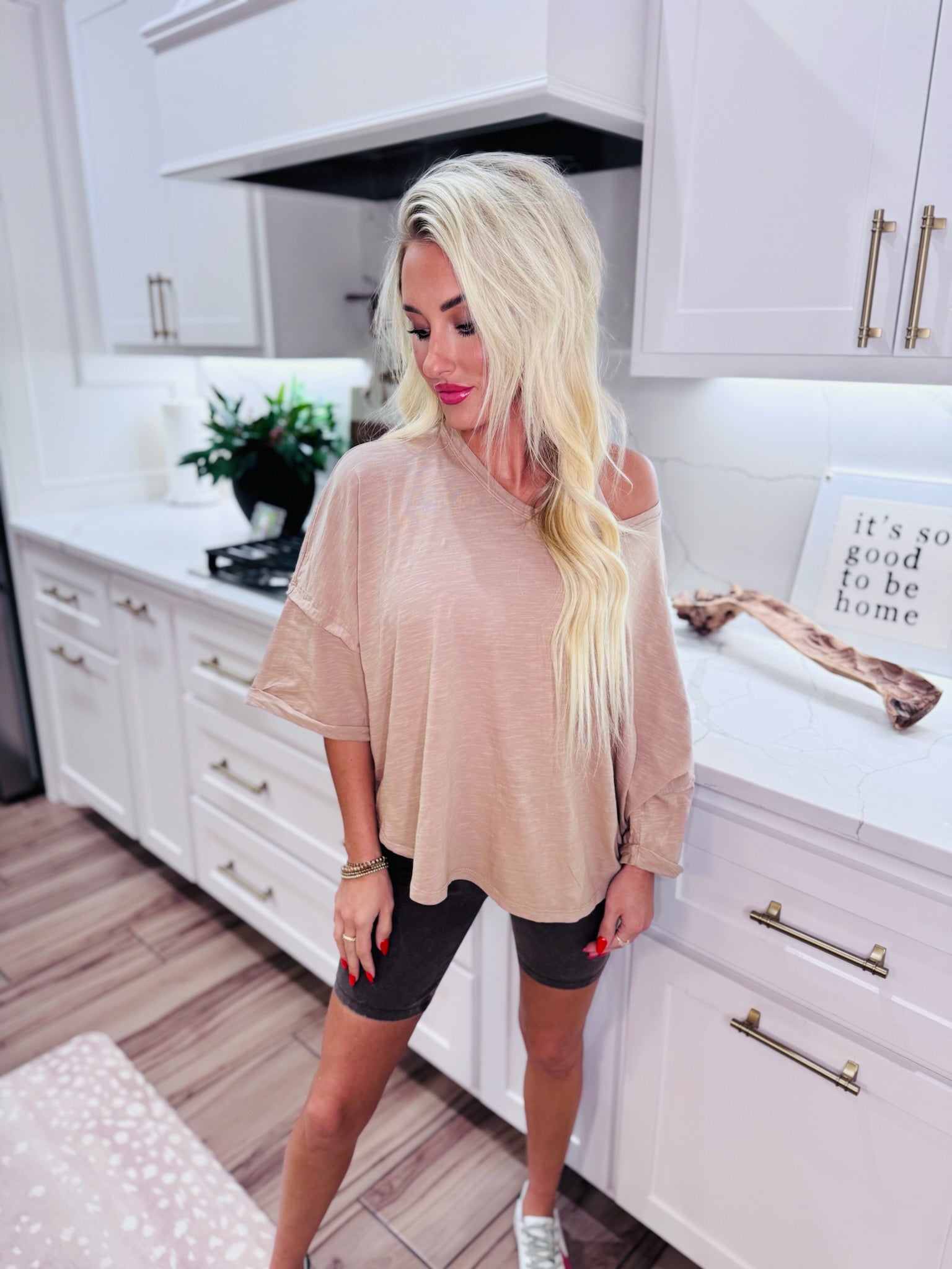Taupe twist back cropped tee on a woman standing in a modern kitchen. The tee has a V-neck and is off-the-shoulder.