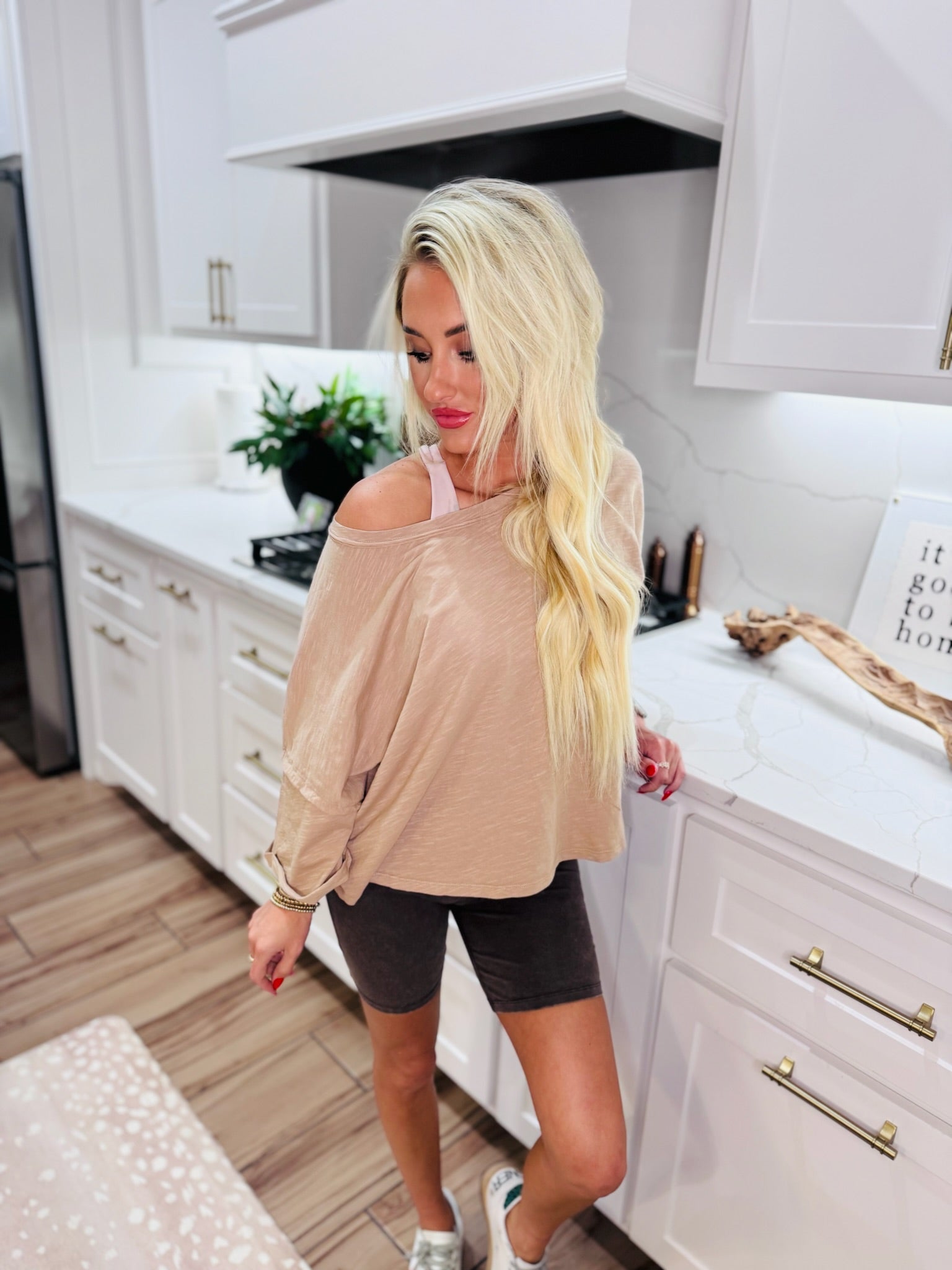 Taupe twist-back cropped tee on blonde woman wearing dark bike shorts and sneakers in a modern kitchen.