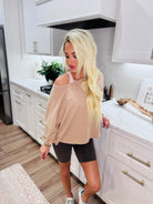 Taupe twist-back cropped tee on blonde woman wearing dark bike shorts and sneakers in a modern kitchen.