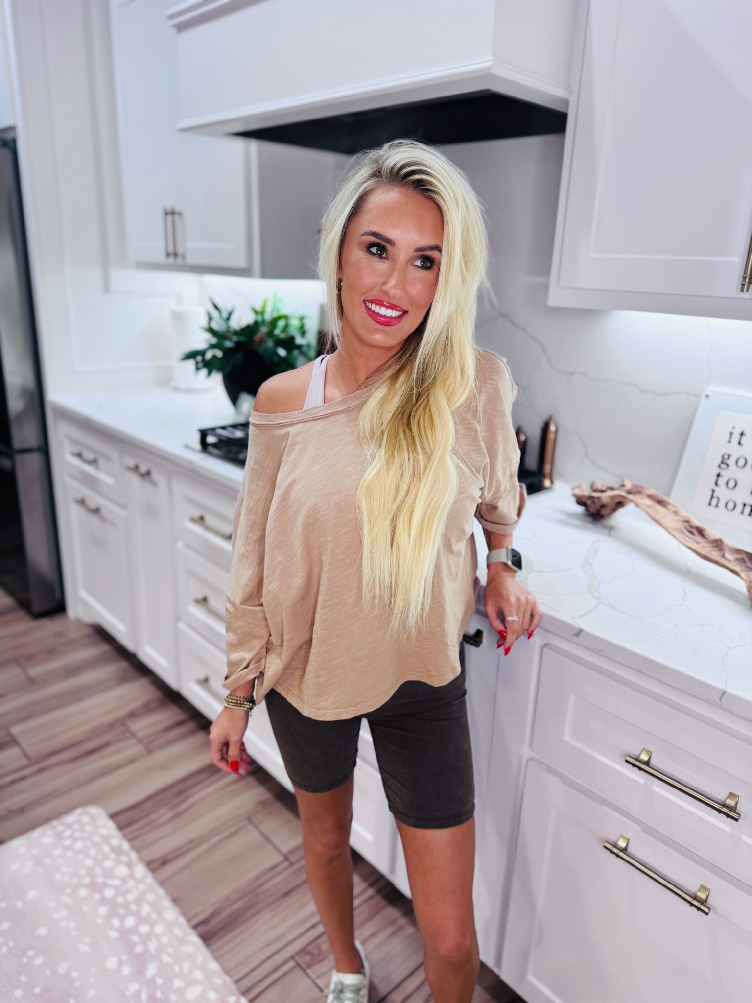 Woman wearing a taupe twist back cropped tee and dark biker shorts, standing in a kitchen.