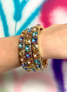 Three stacked gold bracelets adorned with colorful cushion-cut crystals in blue, purple, pink, and amber tones.