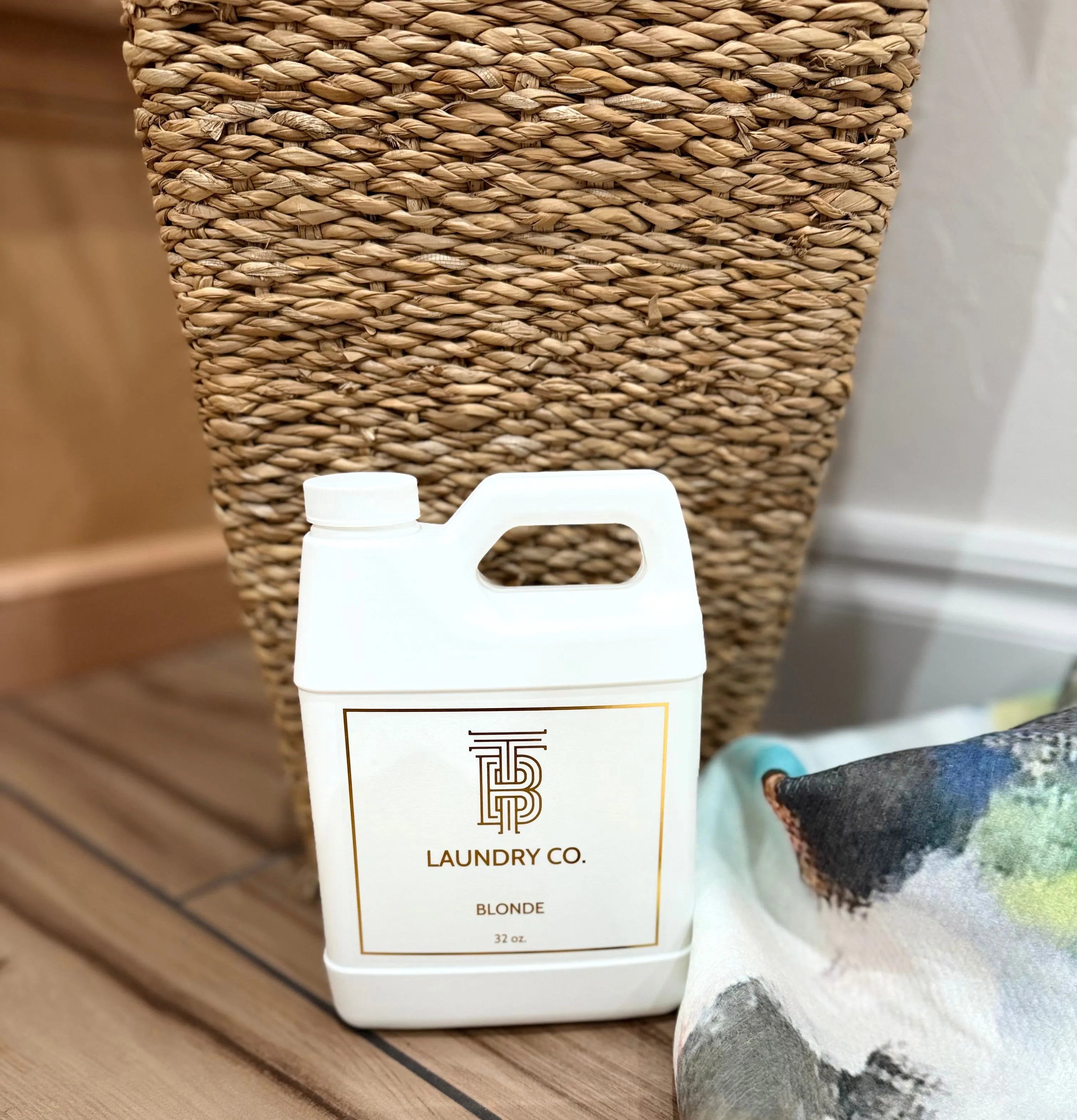 Thomas Blonde Laundry Co. 32oz scented laundry detergent in white jug with gold label, next to a wicker basket and colorful
