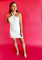 White tweed halter mini dress with pearl trim, worn by a model against a pink backdrop.