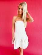 Woman in a white strapless mini dress with tiered ruffles at the hem, posing against a pink background. Ideal for