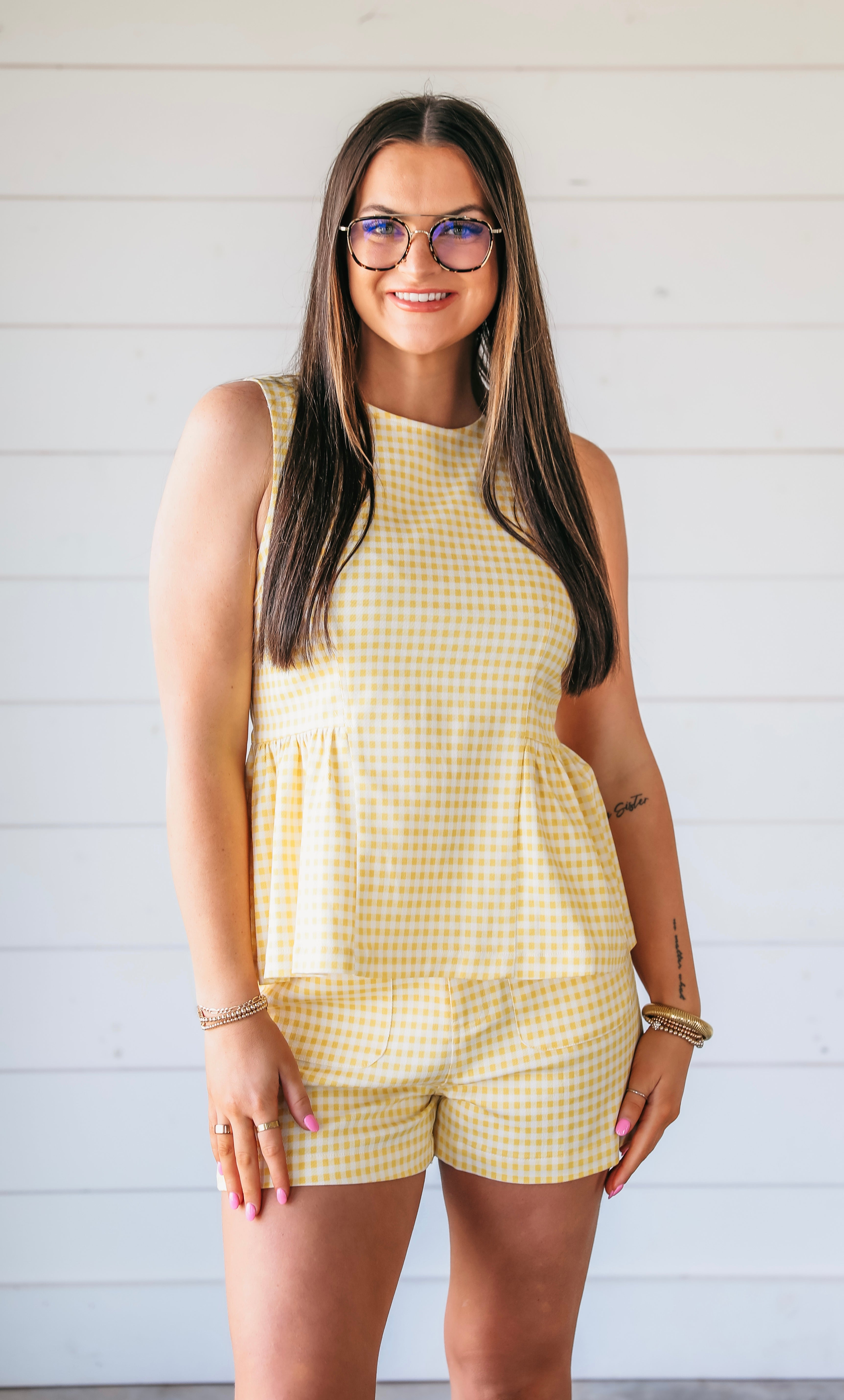 Woman wearing a yellow gingham peplum top and shorts set. She has on glasses and bracelets.